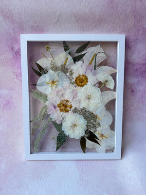 Pressed white flowers and greenery in a white floating frame, shown upright.