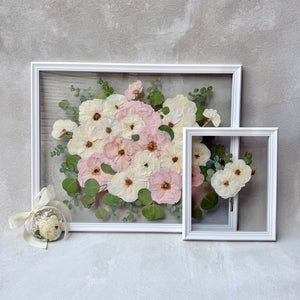 Pressed blush and cream flowers with greenery in a white floating frame, with a matching smaller frame and an ornament.