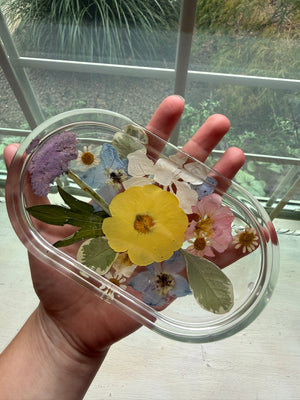 Clear oval resin ring dish with preserved yellow, blue and white flowers, held in a hand.
