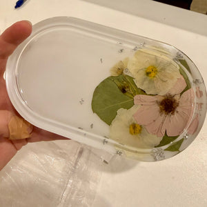 Frosted oval resin ring dish with preserved pale yellow, pink and white flowers and a green leaf, held in a hand.