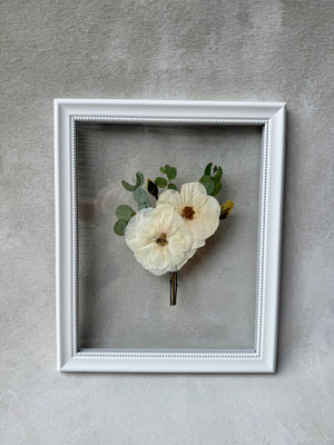 White boutonniere frame with a pressed white flower and greenery, shown upright.