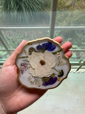 Organic-shaped resin coaster with a gold edge and preserved white, blue and purple flowers, held in a hand.
