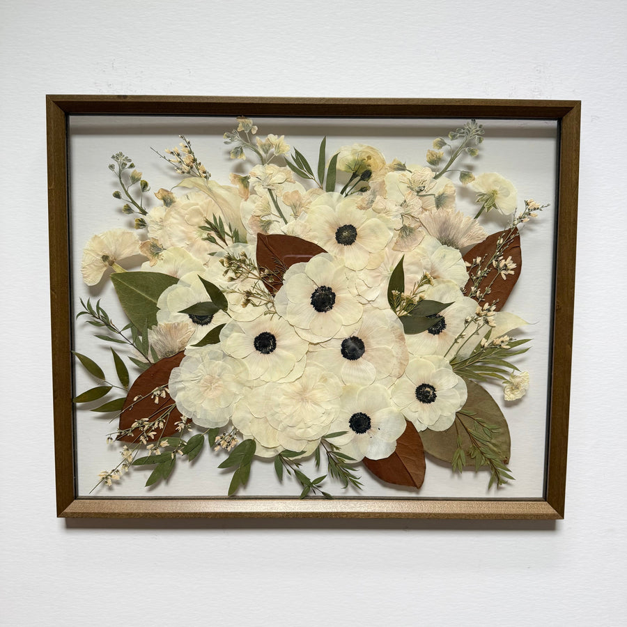 Pressed white flowers with dark centers, greenery and bronze leaves in a dark wood frame.