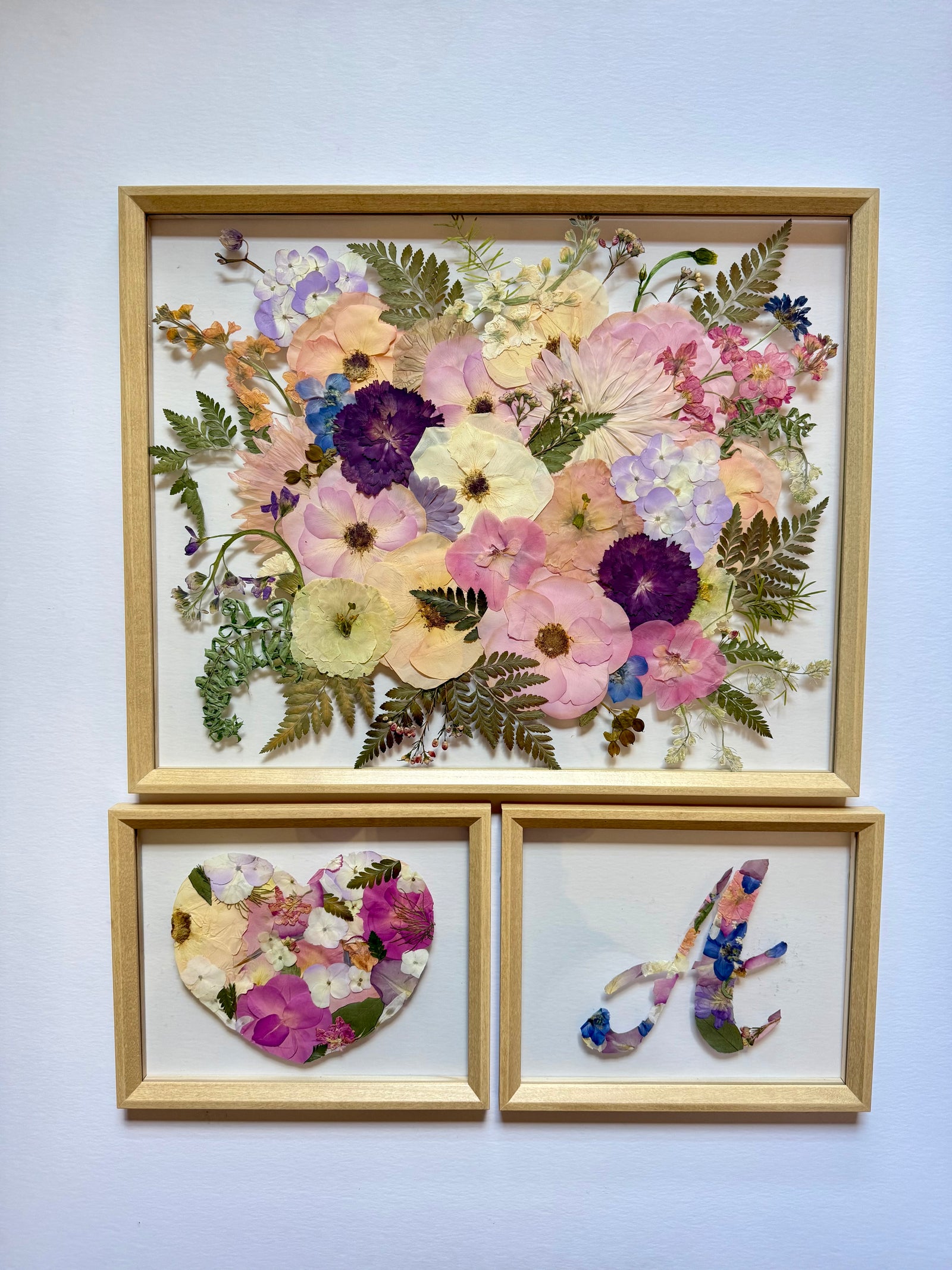 Pressed pink, purple and peach flowers in a light wood frame, with two small frames: a heart and a monogram letter.