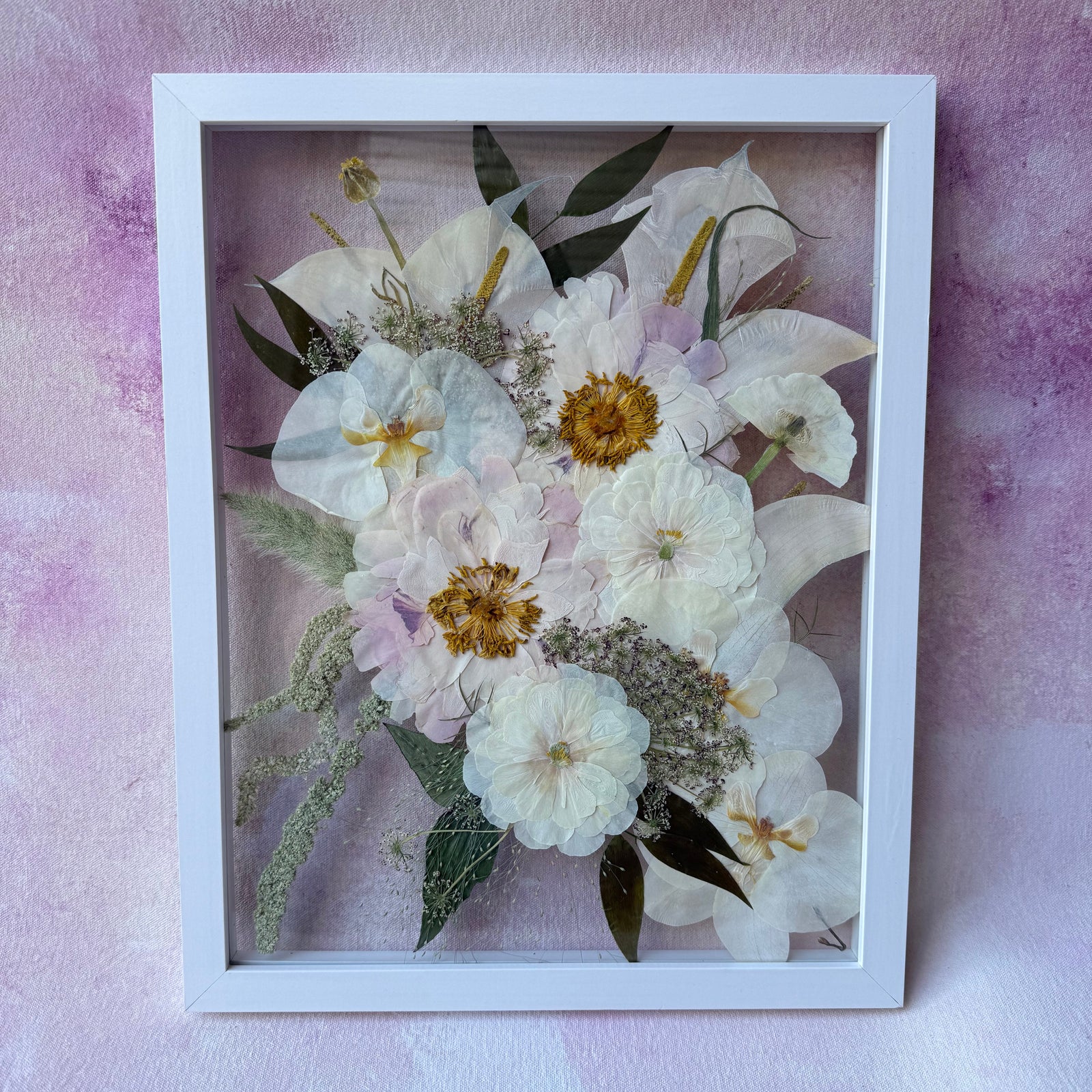Pressed white and cream flowers with dark leaves in a white floating frame, shown upright.