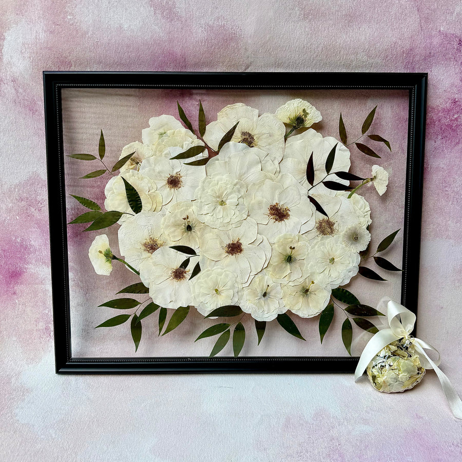 Pressed white and cream flowers with green leaves in a black floating frame, shown with a flower ornament.