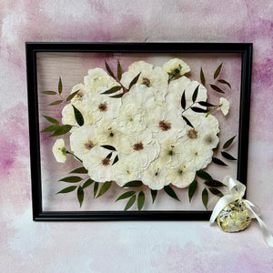 Pressed white and cream flowers with green leaves in a black floating frame, shown with a flower ornament.