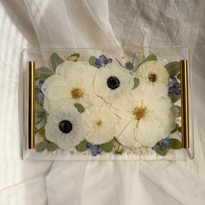 Clear resin rectangle tray with gold handles and preserved white flowers with dark centers and small blue flowers.